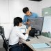 Two young professionals collaborating at a desk with laptops and monitors in a modern office setting.