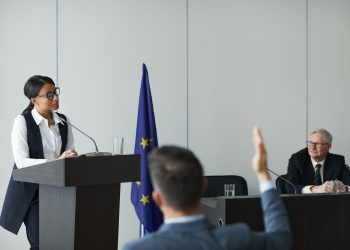 A woman speaking at a European Union conference while delegates listen attentively.