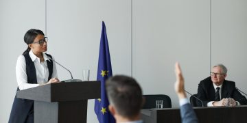 A woman speaking at a European Union conference while delegates listen attentively.