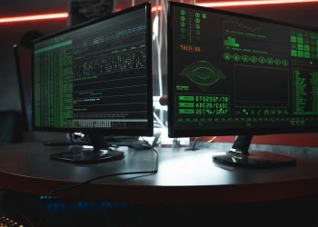 Close-up of dual computer monitors with green coding interfaces in a dark room, highlighting cyber security themes.