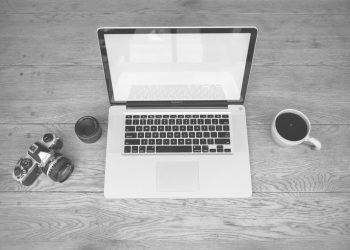 Monochrome image of a laptop, camera, lens, and coffee cup on a wooden desk