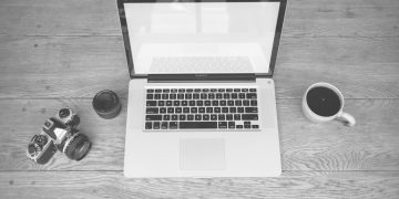 Monochrome image of a laptop, camera, lens, and coffee cup on a wooden desk
