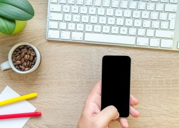 Overhead view of a hand holding smartphone near keyboard, mug of coffee beans, and plant on wooden desk.