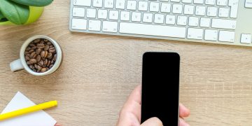 Overhead view of a hand holding smartphone near keyboard, mug of coffee beans, and plant on wooden desk.