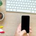 Overhead view of a hand holding smartphone near keyboard, mug of coffee beans, and plant on wooden desk.