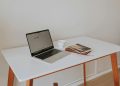 A minimalistic workspace featuring a laptop, coffee cup, and books on a white desk with wooden legs.
