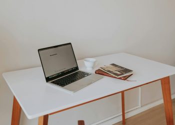 A minimalistic workspace featuring a laptop, coffee cup, and books on a white desk with wooden legs.
