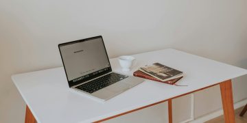 A minimalistic workspace featuring a laptop, coffee cup, and books on a white desk with wooden legs.