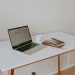 A minimalistic workspace featuring a laptop, coffee cup, and books on a white desk with wooden legs.
