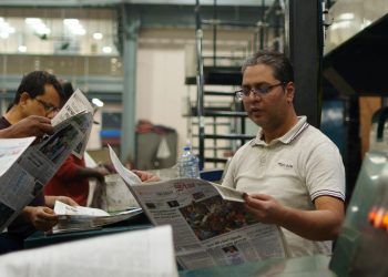 Workers at a newspaper printing press reading freshly printed copies. Focus on a man holding a newspaper.