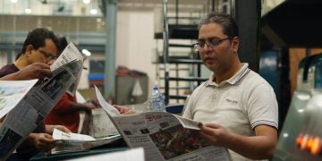 Workers at a newspaper printing press reading freshly printed copies. Focus on a man holding a newspaper.
