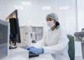 Scientist using a computer in a laboratory with protective gear including gloves, mask, and hair net.