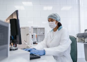 Scientist using a computer in a laboratory with protective gear including gloves, mask, and hair net.
