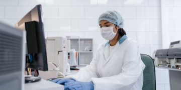 Scientist using a computer in a laboratory with protective gear including gloves, mask, and hair net.