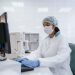 Scientist using a computer in a laboratory with protective gear including gloves, mask, and hair net.
