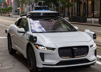 A white autonomous vehicle navigating a city street, reflecting urban architecture in daylight.