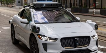 A white autonomous vehicle navigating a city street, reflecting urban architecture in daylight.