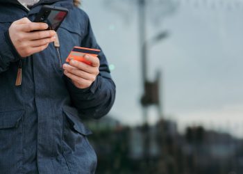 Crop anonymous man in warm clothes holding credit card and browsing smartphone while standing on street in daytime