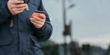 Crop anonymous man in warm clothes holding credit card and browsing smartphone while standing on street in daytime