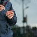 Crop anonymous man in warm clothes holding credit card and browsing smartphone while standing on street in daytime