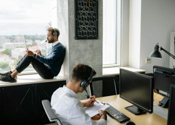 Two call center agents working in a modern office, one seated at a computer and another using a tablet by the window.