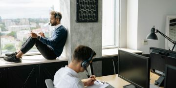 Two call center agents working in a modern office, one seated at a computer and another using a tablet by the window.