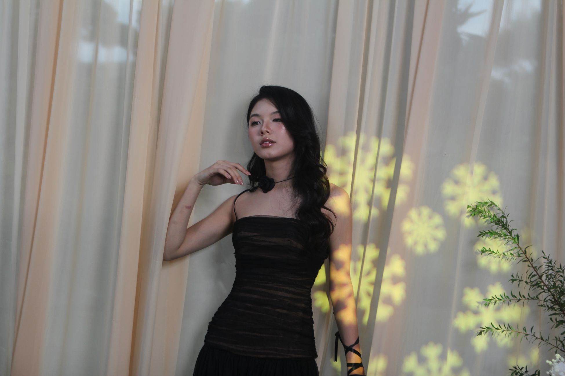 Elegant woman in black dress posing against curtain indoors with artistic light patterns.