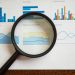 A magnifying glass focuses on various business charts and graphs on paper.
