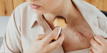 Close-up of a woman using a brush to cover a chest scar with makeup.