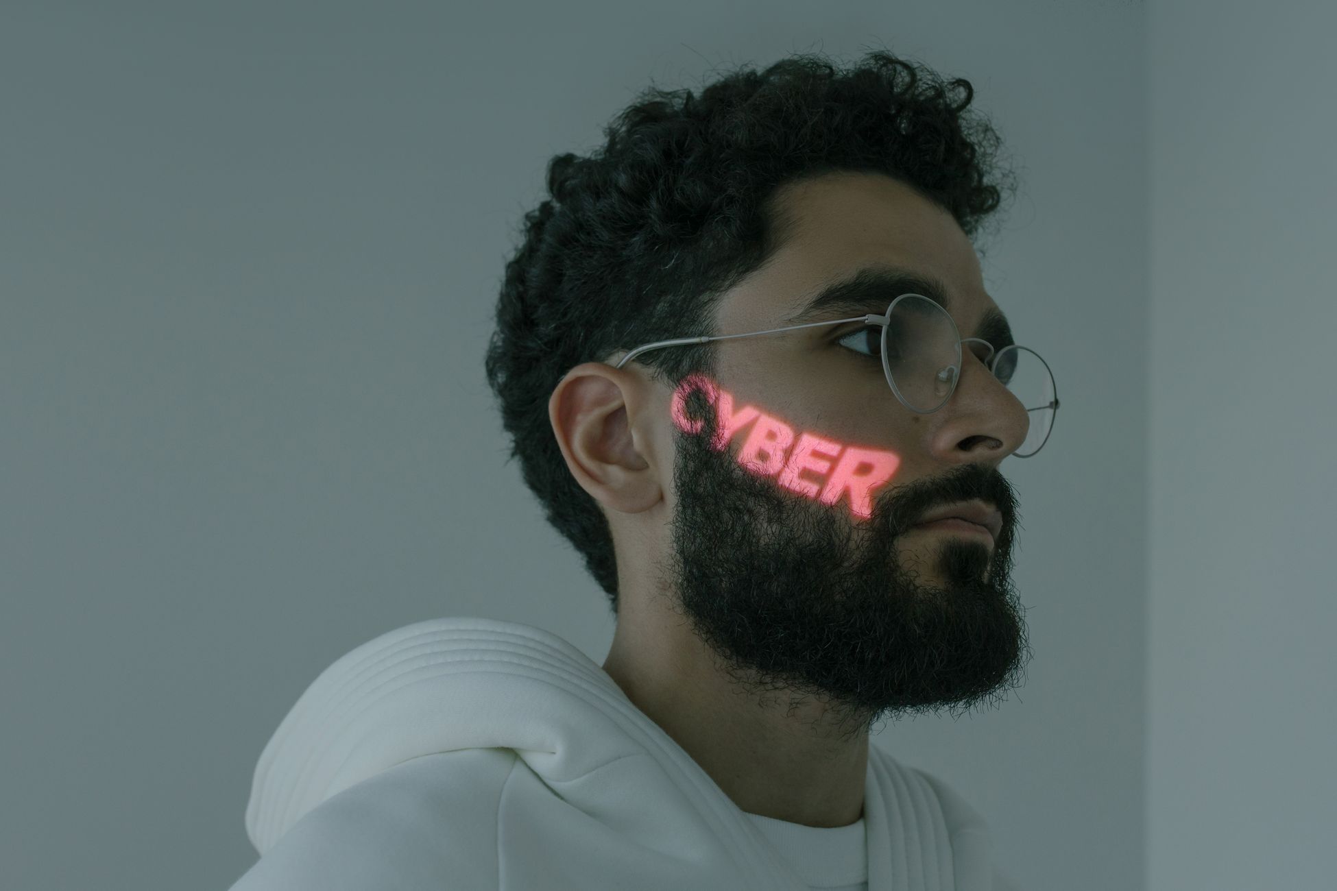 Bearded man with cyber text projection, wearing eyeglasses and white hoodie.