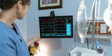 A nurse in blue scrubs examines a medical monitor displaying vital signs in a hospital setting.