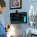 A nurse in blue scrubs examines a medical monitor displaying vital signs in a hospital setting.