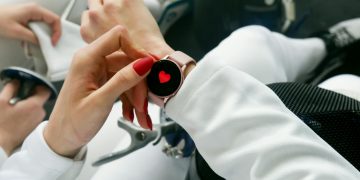 Close-up of a woman's hand with red nails interacting with a smartwatch displaying a heart icon.