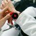 Close-up of a woman's hand with red nails interacting with a smartwatch displaying a heart icon.