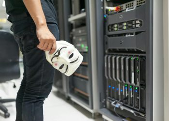 Person holding anonymous mask near servers, hinting at cybersecurity and hacking themes.