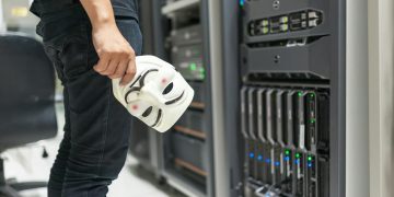 Person holding anonymous mask near servers, hinting at cybersecurity and hacking themes.