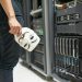 Person holding anonymous mask near servers, hinting at cybersecurity and hacking themes.