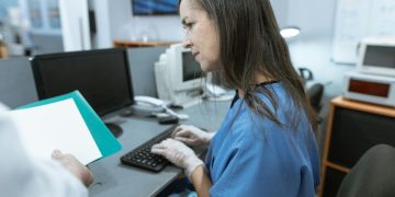 Nurse in scrubs typing on a keyboard at a medical workstation.