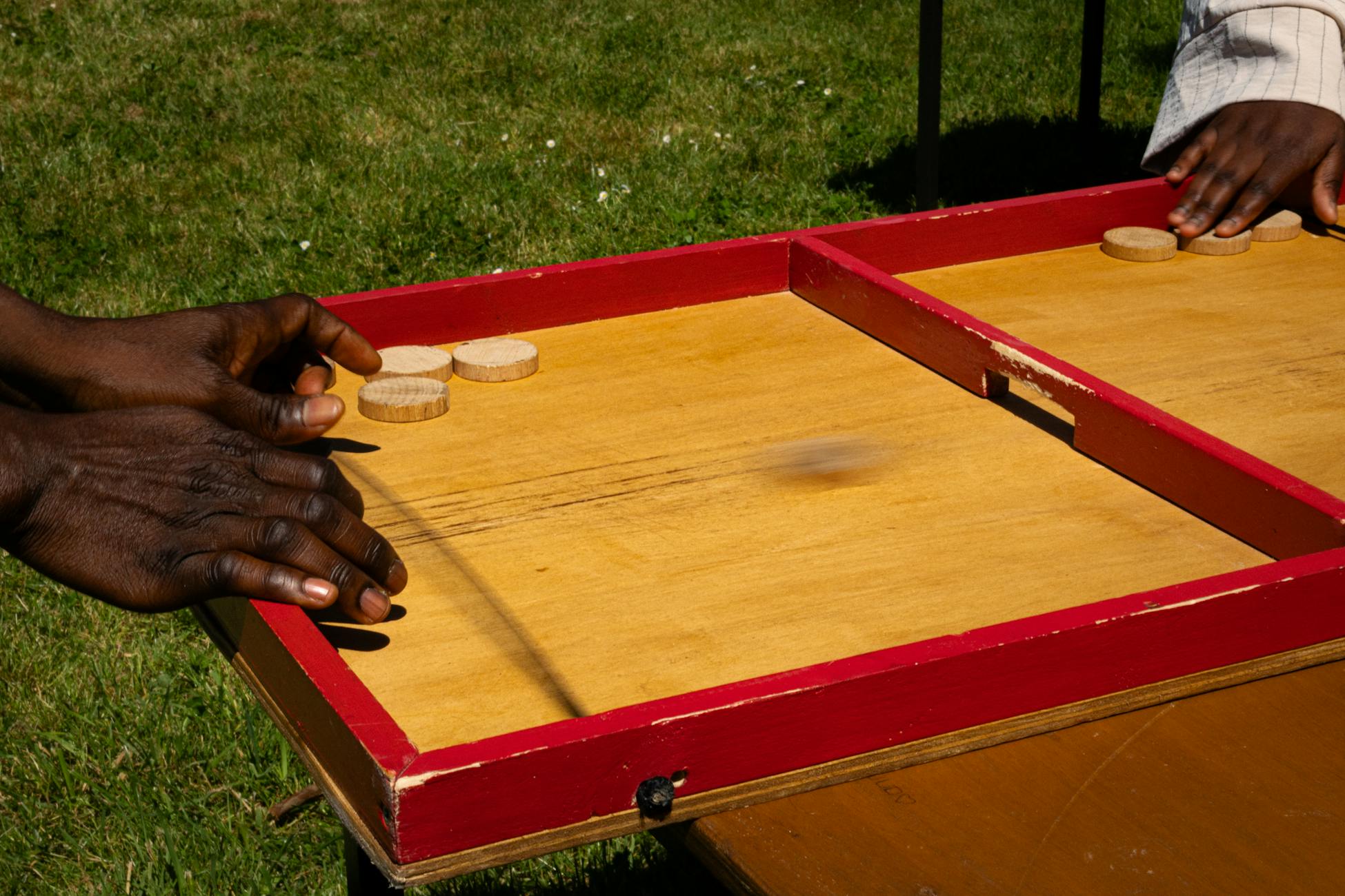 Hands playing a traditional wooden board game outdoors on a sunny day.