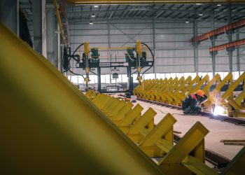 A worker welding metal in an industrial warehouse with heavy machinery.