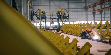 A worker welding metal in an industrial warehouse with heavy machinery.