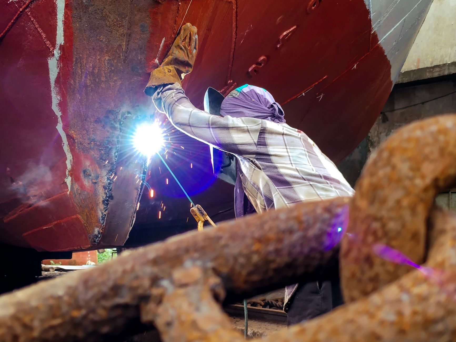 A dedicated worker performs welding on a ship's hull in a shipyard, showcasing industrial skill.