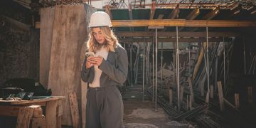 Young female engineer with hard hat checking phone at construction site.
