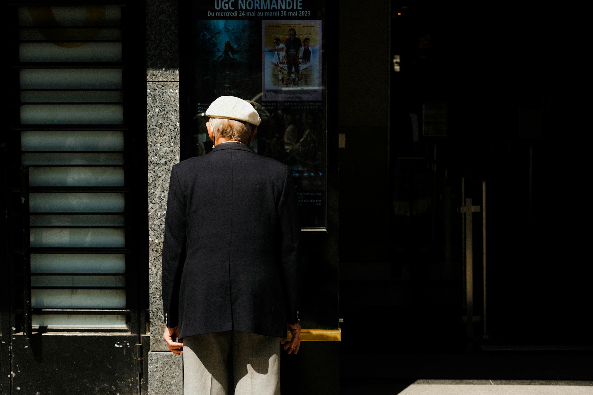 Elderly man in classic attire viewing movie posters outside a cinema.