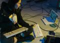A young woman in a dark room surrounded by computers and cables, eating and typing on keyboards.
