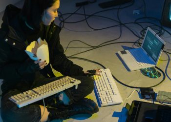 A young woman in a dark room surrounded by computers and cables, eating and typing on keyboards.
