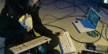 A young woman in a dark room surrounded by computers and cables, eating and typing on keyboards.