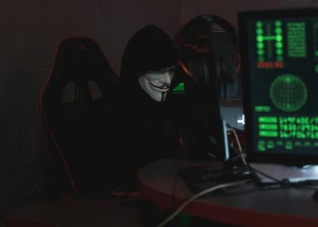 An anonymous hacker wearing a Guy Fawkes mask sits at a computer in a dimly lit room, engaged in cyber activities.