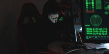 An anonymous hacker wearing a Guy Fawkes mask sits at a computer in a dimly lit room, engaged in cyber activities.