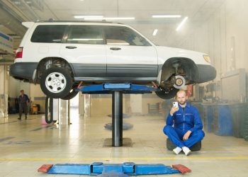 Mechanic in blue coverall using smartphone beside vehicle on lift in repair workshop.